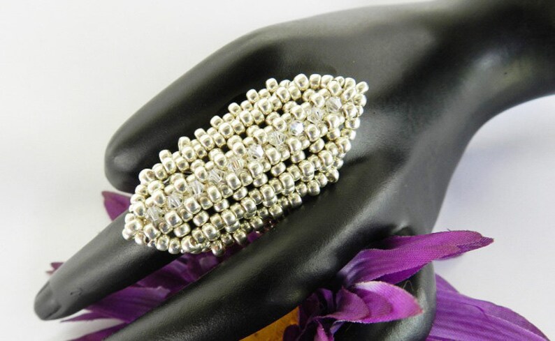 May include: A silver and clear crystal beaded ring. The ring is oval shaped and has a textured design. The ring is on a black hand model.