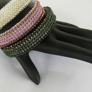 May include: Three beaded bracelets stacked on a black hand-shaped display. The top bracelet is white, the middle bracelet is pink, and the bottom bracelet is green.