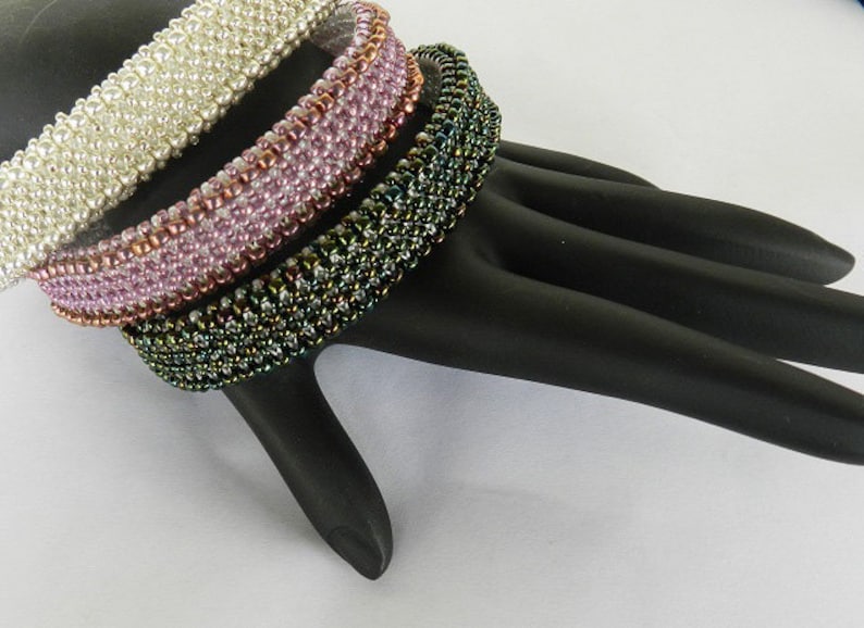May include: Three beaded bracelets stacked on a black hand-shaped display. The top bracelet is white, the middle bracelet is pink, and the bottom bracelet is green.