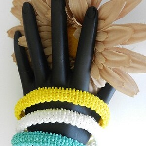 May include: Three beaded bracelets stacked on a black hand model. The bracelets are yellow, white, and turquoise.