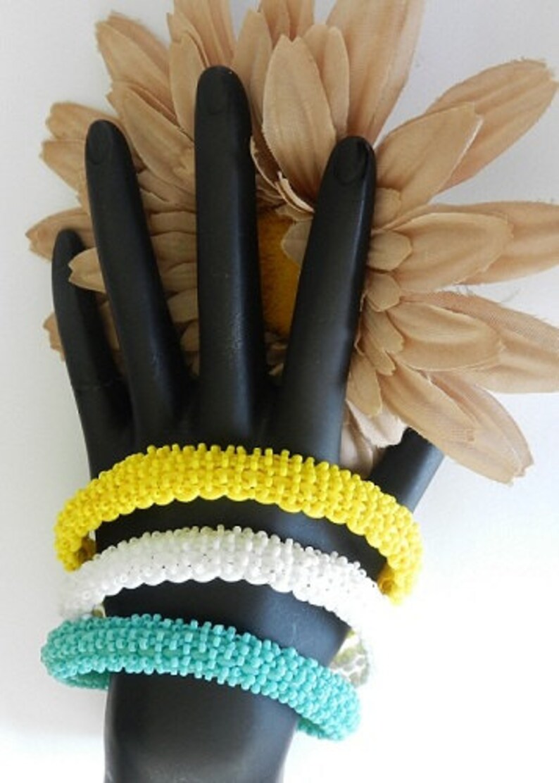 May include: Three beaded bracelets stacked on a black hand model. The bracelets are yellow, white, and turquoise.