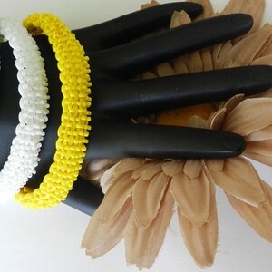 May include: Three beaded bracelets in turquoise, white, and yellow. The bracelets are stacked on a black hand-shaped display stand.