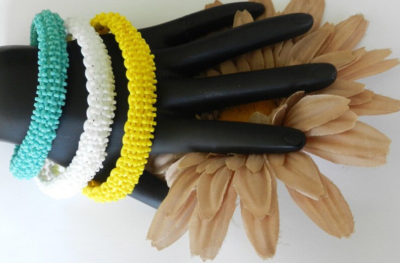May include: Three beaded bracelets in turquoise, white, and yellow. The bracelets are stacked on a black hand-shaped display stand.