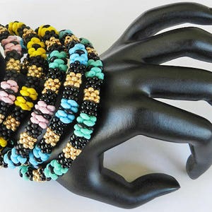 Op de afbeelding: Een set van vijf kleurrijke kralenarmbanden, met zwarte, gele, roze, blauwe en turquoise kralen. De armbanden zijn op elkaar gestapeld en worden gedragen op een zwarte mannequinhand.