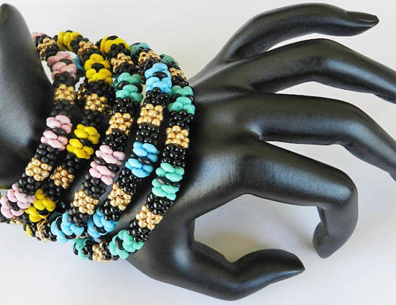 Op de afbeelding: Een set van vijf kleurrijke kralenarmbanden, met zwarte, gele, roze, blauwe en turquoise kralen. De armbanden zijn op elkaar gestapeld en worden gedragen op een zwarte mannequinhand.