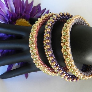 May include: Three beaded bracelets in shades of green, purple, and gold. The bracelets are stacked on a black mannequin hand.