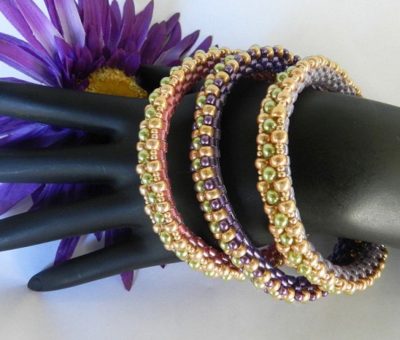 May include: Three beaded bracelets in shades of green, purple, and gold. The bracelets are stacked on a black mannequin hand.