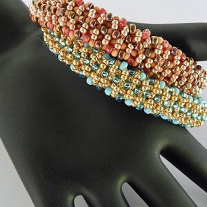 Puede incluir: Tres pulseras de cuentas apiladas en una mano negra. La pulsera superior es roja, naranja y dorada. La pulsera del medio es dorada y turquesa. La pulsera inferior es dorada y turquesa.