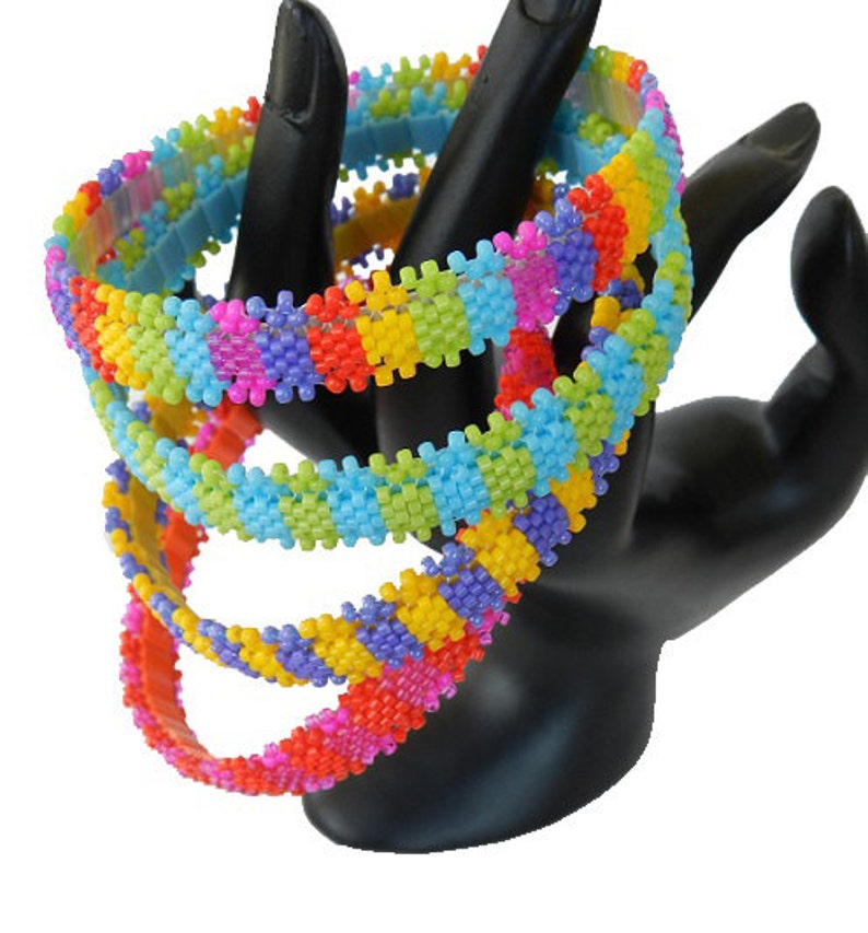 May include: Four colorful beaded bracelets stacked on a black display stand. The bracelets are made of small, square beads in shades of pink, purple, yellow, orange, green, and blue.