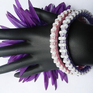 May include: Three beaded bracelets in shades of purple, pink, and white. The bracelets are stacked on a black mannequin hand.