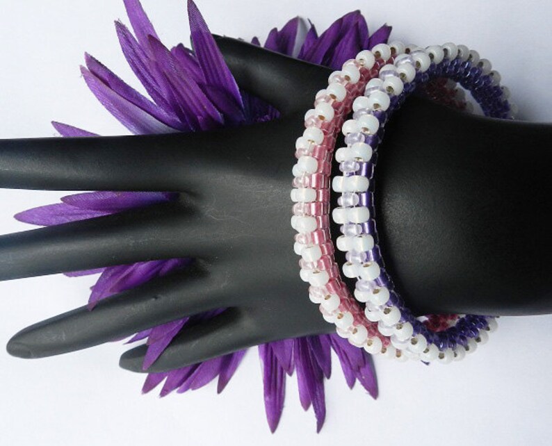 May include: Three beaded bracelets in shades of purple, pink, and white. The bracelets are stacked on a black mannequin hand.