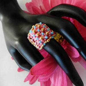 K&ouml;nnte beinhalten: Ein quadratischer Ring aus rosa, blauen und goldenen Perlen. Der Ring befindet sich auf einem schwarzen Handmodell.