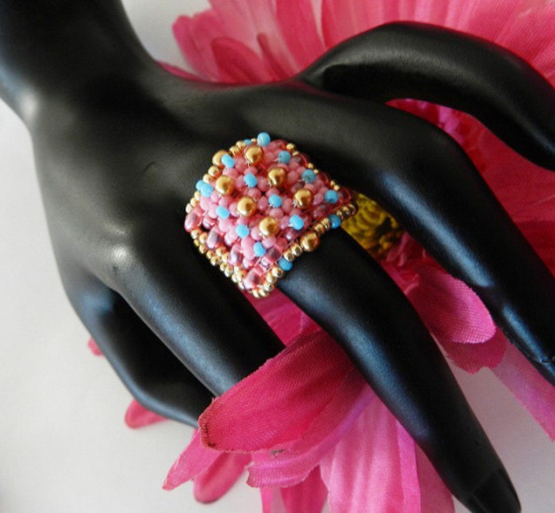 K&ouml;nnte beinhalten: Ein quadratischer Ring aus rosa, blauen und goldenen Perlen. Der Ring befindet sich auf einem schwarzen Handmodell.