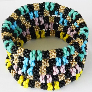 Op de afbeelding: Meerkleurig kralenarmband met zwarte, turquoise, gele en roze kralen. De armband is gemaakt van meerdere rijen kralen en heeft een geweven patroon.