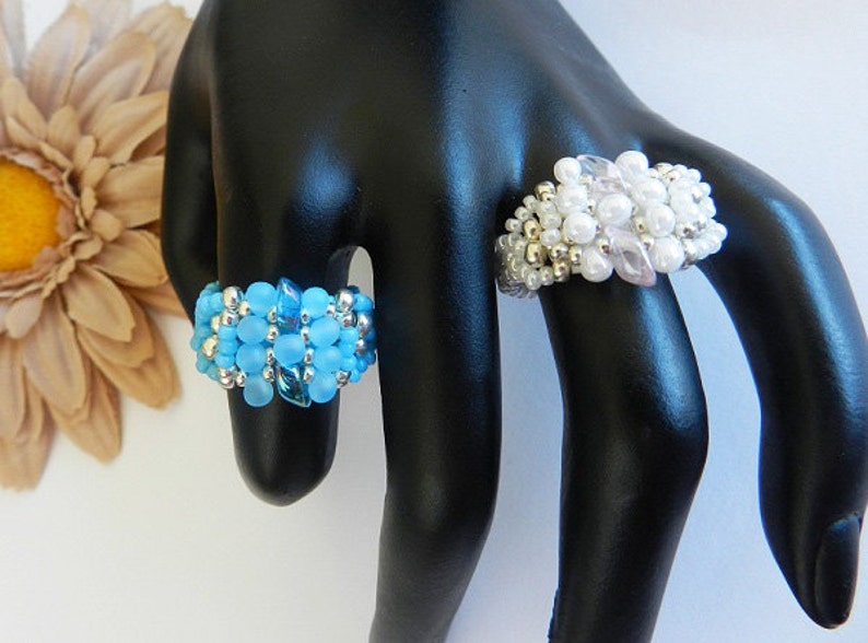 May include: Two beaded rings on a black ring display stand. The ring on the left is blue and silver. The ring on the right is white and silver.