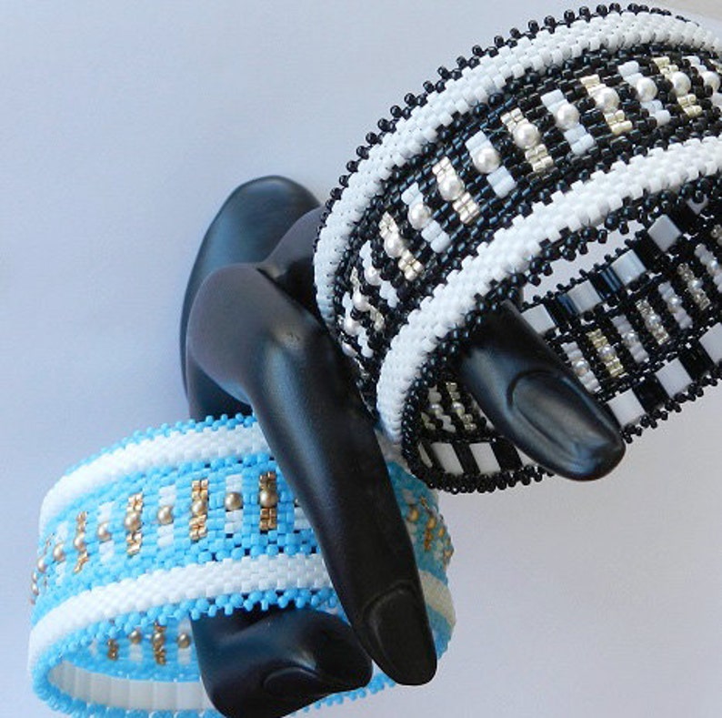 Op de afbeelding: Twee kralenarmbanden, een zwart-wit, de andere blauw, wit en goud, op een zwarte mannequinhand.