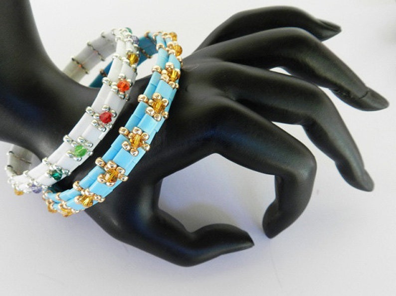 May include: Three beaded bracelets stacked on a black mannequin hand. The top bracelet is white with silver and multi-colored beads. The middle bracelet is light blue with gold and white beads. The bottom bracelet is light blue with gold beads.