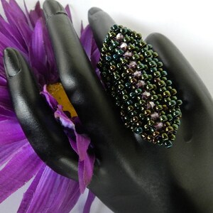 May include: A dark green and purple beaded ring with iridescent beads. The ring is on a black hand model.
