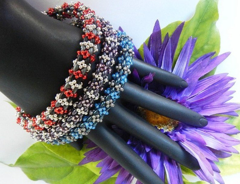 May include: Four beaded bracelets stacked on a black mannequin hand. The bracelets are made of silver, red, blue, and purple beads. The bracelets are on a background of a purple flower.