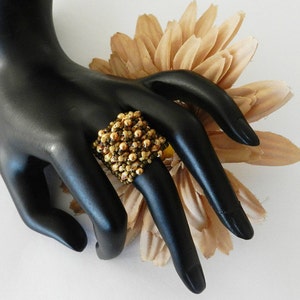 K&ouml;nnte beinhalten: Ein quadratischer Ring aus braunen und goldenen Perlen. Der Ring befindet sich an einer schwarzen Mannequin-Hand.