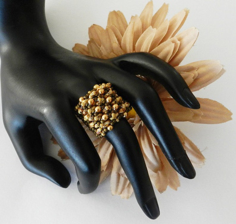 K&ouml;nnte beinhalten: Ein quadratischer Ring aus braunen und goldenen Perlen. Der Ring befindet sich an einer schwarzen Mannequin-Hand.