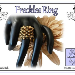 May include: A gold and brown beaded ring, called "Freckles Ring", is shown on a black hand model. The ring is made with square stitch beading. The image is titled "Freckles Ring" and includes the text "DebbiegerDesigns" and "Debbie van Tonder".