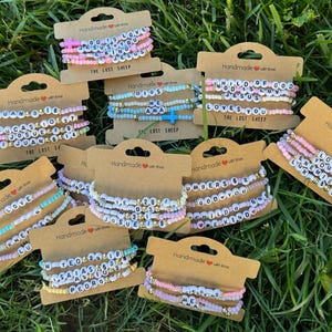 May include: A collection of handmade beaded bracelets in various pastel colors, displayed on brown cardstock. Each bracelet features white letter beads spelling out words and phrases, with gold accent beads. The text "THE LOST SHEEP" is printed on the cards.