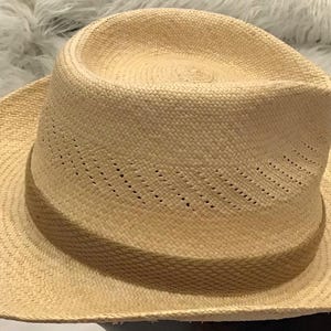 Puede incluir: Un sombrero fedora beige claro con una textura tejida. El sombrero presenta una corona estructurada y un ala ancha. El sombrero tiene peque&ntilde;os orificios de ventilaci&oacute;n a lo largo de la corona. El sombrero est&aacute; hecho de fibra natural.