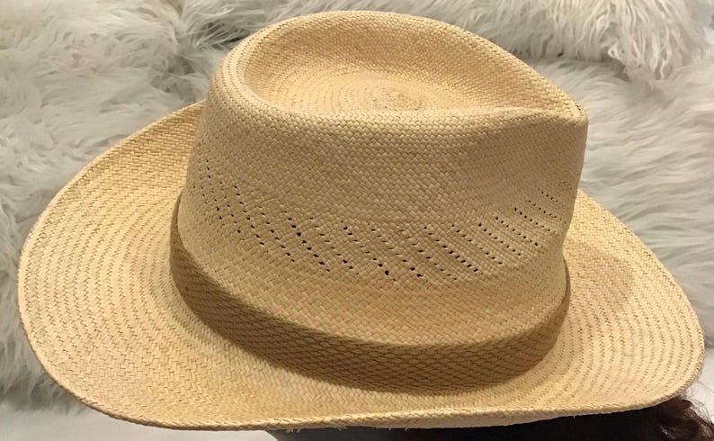 Puede incluir: Un sombrero fedora beige claro con una textura tejida. El sombrero presenta una corona estructurada y un ala ancha. El sombrero tiene peque&ntilde;os orificios de ventilaci&oacute;n a lo largo de la corona. El sombrero est&aacute; hecho de fibra natural.