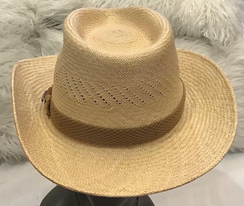 Puede incluir: Un sombrero fedora de paja de color beige claro con una banda marr&oacute;n. El sombrero tiene un dise&ntilde;o perforado en la corona y un ala ancha. El sombrero se muestra sobre una superficie oscura.