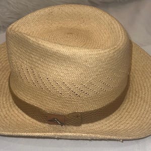Puede incluir: Sombrero de paja de color beige claro con ala ancha y copa ligeramente hundida. El sombrero presenta una banda de cuero marr&oacute;n y un cierre met&aacute;lico decorativo. La textura tejida del sombrero es visible.