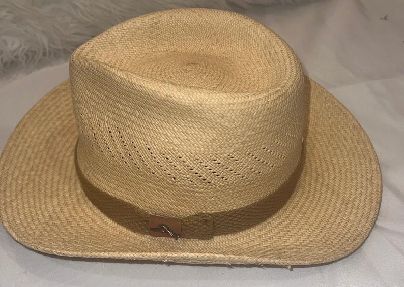 Puede incluir: Sombrero de paja de color beige claro con ala ancha y copa ligeramente hundida. El sombrero presenta una banda de cuero marr&oacute;n y un cierre met&aacute;lico decorativo. La textura tejida del sombrero es visible.
