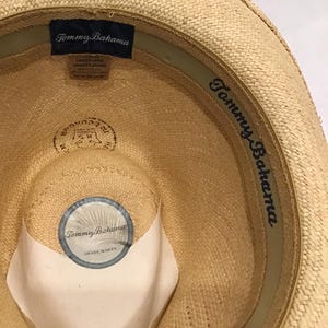 Puede incluir: Sombrero de paja Tommy Bahama de color beige claro. El interior presenta una etiqueta azul marino con el nombre de la marca y la talla, y una pegatina circular que dice "Tommy Bahama Shade Maker". El sombrero tiene una textura tejida y una banda verde.