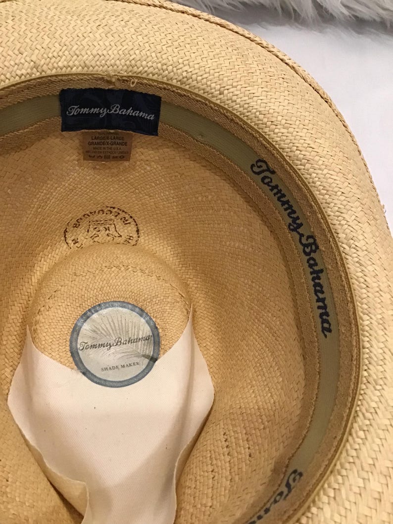 Puede incluir: Sombrero de paja Tommy Bahama de color beige claro. El interior presenta una etiqueta azul marino con el nombre de la marca y la talla, y una pegatina circular que dice "Tommy Bahama Shade Maker". El sombrero tiene una textura tejida y una banda verde.