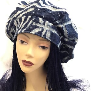 May include: A navy blue and gray patterned beret. The beret features a repeating zebra-like pattern in navy blue and gray. The hat is displayed on a mannequin head with dark blue braids and dark lipstick.