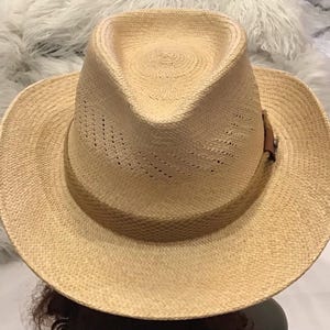 Puede incluir: Sombrero fedora de color beige claro hecho de paja tejida. El sombrero presenta una banda de cuero marr&oacute;n con un peque&ntilde;o elemento decorativo. La corona tiene una ligera hendidura y el ala est&aacute; ligeramente curvada. El sombrero est&aacute; dise&ntilde;ado para la protecci&oacute;n solar.