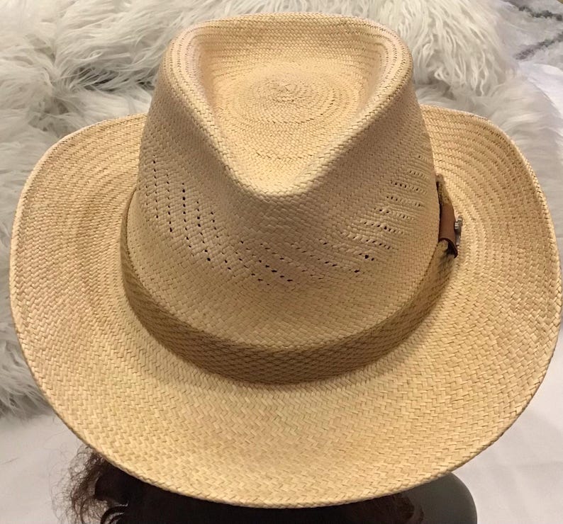 Puede incluir: Sombrero fedora de color beige claro hecho de paja tejida. El sombrero presenta una banda de cuero marr&oacute;n con un peque&ntilde;o elemento decorativo. La corona tiene una ligera hendidura y el ala est&aacute; ligeramente curvada. El sombrero est&aacute; dise&ntilde;ado para la protecci&oacute;n solar.