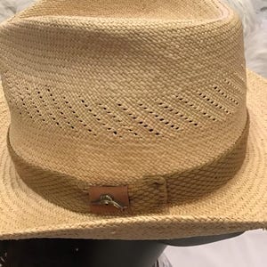 Puede incluir: Sombrero fedora de paja tejida con una banda marr&oacute;n y un peque&ntilde;o emblema met&aacute;lico. El sombrero tiene un dise&ntilde;o cl&aacute;sico con una corona ligeramente hundida y un ala ancha. El sombrero es de color marr&oacute;n claro.