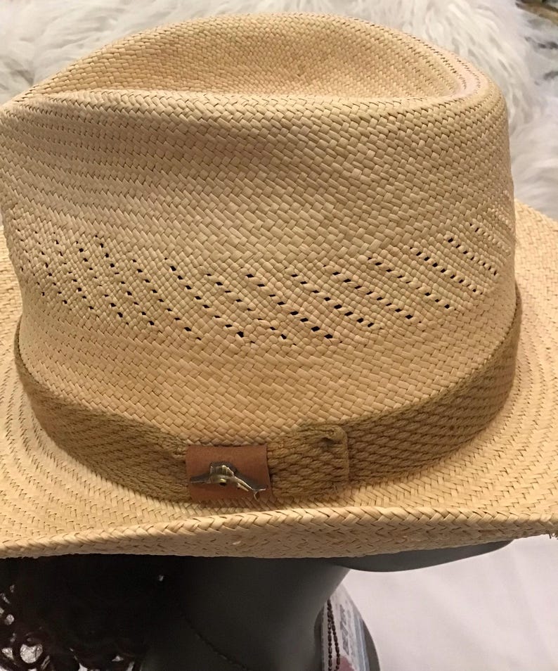 Puede incluir: Sombrero fedora de paja tejida con una banda marr&oacute;n y un peque&ntilde;o emblema met&aacute;lico. El sombrero tiene un dise&ntilde;o cl&aacute;sico con una corona ligeramente hundida y un ala ancha. El sombrero es de color marr&oacute;n claro.