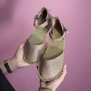 Pode incluir: Um par de sandálias de couro marrom claro com tiras de tornozelo e fivelas douradas. As sandálias têm uma borda costurada e uma sola plana.