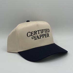 May include: A beige and navy blue baseball cap. The cap features the text "CERTIFIED YAPPER" embroidered in black on the front. The cap has a structured crown and a curved brim.