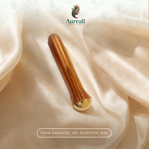Puede incluir: Una herramienta de masaje facial ayurv&eacute;dica de madera con un extremo dorado. La herramienta es marr&oacute;n con un patr&oacute;n de rayas y la marca "Aureali" est&aacute; impresa en el lateral. Tambi&eacute;n es visible el texto "Glow naturally, the Ayurvedic way".