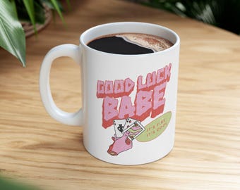 Może przedstawiać: Biały ceramiczny kubek z różowo-zielonym wzorem graficznym z napisem "Good Luck Babe" i "It's Fine It's Cool". Kubek jest wypełniony kawą.