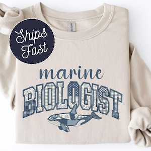 May include: A beige sweatshirt with the words "marine BIOLOGIST" in blue, with a whale graphic. The text and whale design have a patterned texture. A navy blue circle with the words "Ships Fast" is in the upper left corner.