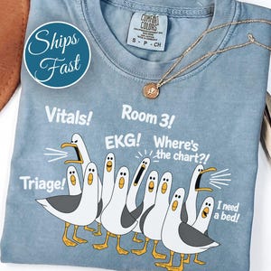 May include: A blue crew neck t-shirt featuring a graphic of multiple seagulls with white text that reads Vitals!, Room 3!, EKG!, Where's the chart?!, Triage!, and I need a bed!. The shirt has a Comfort Colors brand label at the inner neckline.
