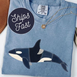 May include: A light blue Comfort Colors t-shirt featuring an appliqued orca whale design in shades of blue, black, and white. A gold necklace with a pendant rests on the shirt. The shirt has the text "Ships Fast" on a navy blue circle.