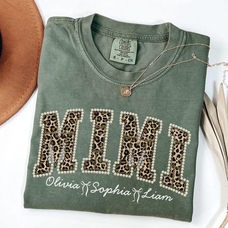 May include: An olive green t-shirt featuring the word "MIMI" in leopard print letters, edged with white pearls. Underneath, the names "Olivia Sophia Liam" are written in script. A gold necklace is placed across the shirt.