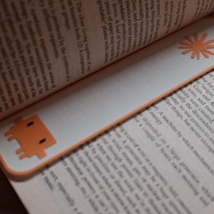 May include: A rectangular white bookmark with an orange border, featuring a pixelated orange character at the bottom and an orange sun-shaped icon with a circular hole at the top, placed between the pages of an open book with printed text.