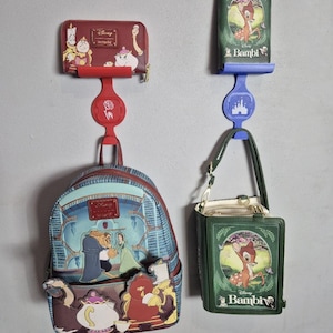 May include: Two Disney-themed bags and wallets are displayed. A red wallet with Beauty and the Beast characters hangs above a matching backpack. A green Bambi-themed purse is displayed next to a matching wallet.