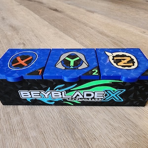 Pode incluir: Uma caixa de armazenamento Beyblade X azul e preta com três compartimentos. Cada compartimento tem um símbolo diferente na tampa: um X vermelho, um Y verde e um Z dourado. A caixa tem o texto "Beyblade X" e "ベイブレードエックス" impresso na frente.